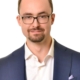 Ein gut gekleideter Mann steht vor einem schlichten weißen Hintergrund. Er hat kurze braune Haare, eine Brille und einen gepflegten Bart. Er trägt einen blauen Anzug über einem frischen weißen Hemd, hält eine weiße Tasse in der Hand und lächelt leicht.