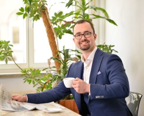 Eine mögliche Beschreibung oder Aufforderung für Ihre Szene könnte lauten: Ein stilvoller Mann namens Alexander, gekleidet in einen eleganten blauen Anzug und mit Brille, sitzt gemütlich an einem Tisch. Er lächelt herzlich, hält eine weiße Kaffeetasse in der Hand, während vor ihm eine Zeitung auf dem Tisch liegt. Üppige grüne Pflanzen und große Fenster im Hintergrund füllen den Raum mit natürlichem Licht und schaffen eine helle und einladende Atmosphäre. Wenn Sie eine Aufforderung zur Bildgenerierung basierend auf dieser Beschreibung wünschen oder Hilfe bei der Verfeinerung (für die Verwendung mit KI/Künstlern) benötigen, lassen Sie es mich wissen!.