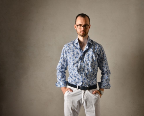 Callboy Alexander steht vor einem schlichten grauen Hintergrund. Er hat kurze Haare und trägt eine Brille. Er ist mit einem blauen Hemd mit Paisleymuster und einer weißen Hose bekleidet. Seine Hände ruhen lässig in den Hosentaschen, während er geradeaus blickt und einen neutralen Blick aufrechterhält.