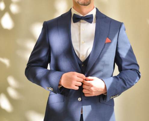 Callboy Alexander steht selbstbewusst vor einem sanft beleuchteten, gesprenkelten Hintergrund. Er trägt einen eleganten blauen Anzug, dazu ein frisches weißes Hemd und eine elegante Fliege. Seine Brille umrahmt scharfe Augen, und sein gepflegter Bart betont seine Kinnpartie. Mit beiden Händen zupft er sorgfältig an seinem Jackett am Revers und strahlt im sanften Sonnenlicht, das die Szenerie umspielt, Gelassenheit und Charme aus.