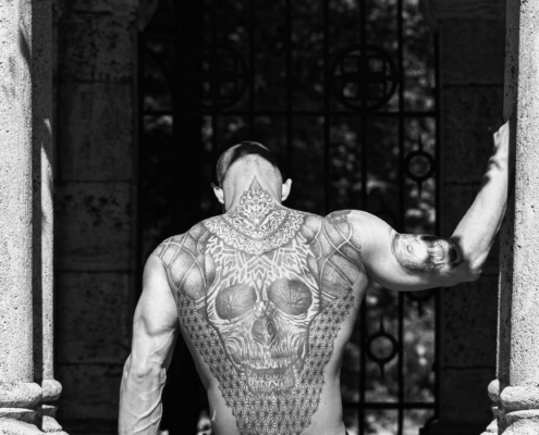 In dieser eindrucksvollen Szene strahlt Callboy Alex eine geheimnisvolle Aura aus, während er mit nacktem Oberkörper zwischen den alten Steinsäulen sitzt. Seine Tattoos, die einen markanten Totenkopf mit komplexen geometrischen Mustern zeigen, erzählen ihre eigene Geschichte. Die historische Kulisse ist voller Charakter, und verwitterte Steine zeugen vom Lauf der Zeit. Die schmiedeeisernen Details sorgen für eine zusätzliche Ebene der Faszination, werfen komplizierte Schatten auf den Boden und verstärken das Gefühl des Mysteriums, das Alex umgibt. Es ist ein Tableau, in dem sich Geschichte und Moderne kreuzen und ein fesselndes Bild entsteht, das sowohl beeindruckend als auch rätselhaft ist.