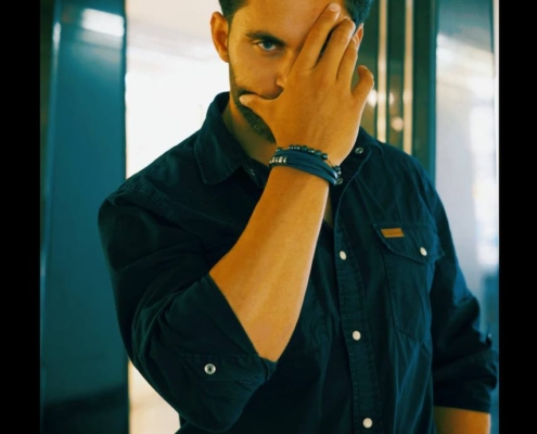 Ein Mann mit dunklem Haar und Bart steht in einem Haus, bekleidet mit einem schwarzen Button-up-Hemd und einer hellen Hose. Er ist als Callboy Matteo bekannt und bedeckt ein Auge mit seiner Hand, wobei er die Kamera mit einem direkten und ernsten Ausdruck fixiert. Die Gesamtstimmung ist intensiv und introspektiv, unterstrichen durch den Kontrast zwischen seiner dunklen Kleidung und dem hellen Hintergrund.