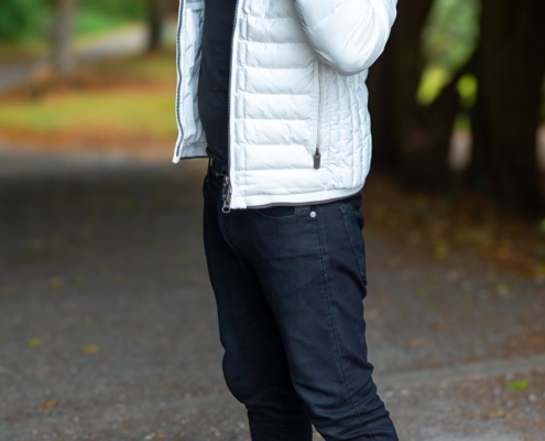 Callboy Kevin steht in einem weißen Puff, einem schwarzen Hemd und Jeans auf einem Kiesweg und fasst sich an den Kragen, während im Hintergrund Bäume stehen.