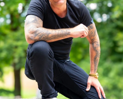 Callboy Kevin, tätowiert und mit kurzen Haaren, kniet in schwarzem Outfit und weißen Turnschuhen im Freien auf einem Stein und lächelt inmitten grüner Bäume.