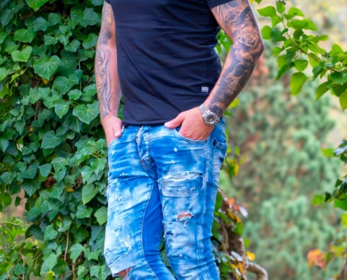 Das von Ihnen beschriebene Bild fängt einen lockeren und entspannten Moment im Freien ein. Der Stil von Callboy Kevin mit seinen kurzen Haaren, Tattoos und seiner Kleidungswahl – schwarzes T-Shirt, verwaschene Bluejeans und weiße Turnschuhe – vermittelt einen modernen und kantigen Look. Das Anlehnen an die efeubewachsene Wand verleiht der Szene einen Hauch von Natur und schafft einen interessanten Kontrast zwischen den urbanen Modeelementen und der natürlichen Kulisse. Seine Hände in den Taschen und sein Seitenblick vermitteln ein zugängliches und doch selbstbewusstes Auftreten.