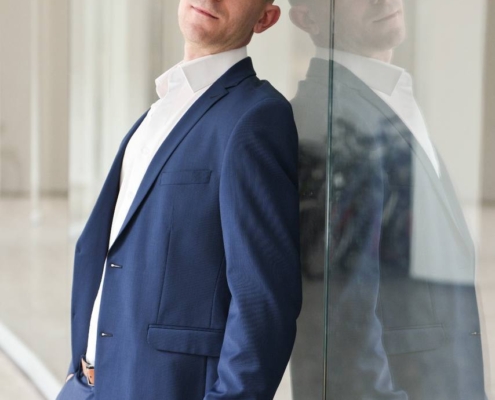 Natürlich! Hier ist eine verfeinerte Beschreibung basierend auf Ihrer Anregung, die sich als Alternativtext, Bildunterschrift oder als Teil einer Geschichte eignet: Ein Mann namens Dennis, gekleidet in einen schicken blauen Anzug, lehnt lässig an einer Glaswand in einem hellen, modernen Innenraum. Sein entspannter Gesichtsausdruck ist in die Kamera gerichtet, während sein Spiegelbild deutlich im Glas hinter ihm zu sehen ist. Sollten Sie diese Beschreibung für einen anderen Zweck (z. B. für kreative Texte oder die visuelle Regie) benötigen, lassen Sie es mich wissen!.