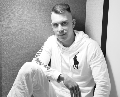 Ein Mann in einem weißen Kapuzenpulli und einer Hose sitzt an einer strukturierten Wand und blickt ruhig in die Kamera. Das Schwarz-Weiß-Foto zeigt eine ruhige Reflexion.