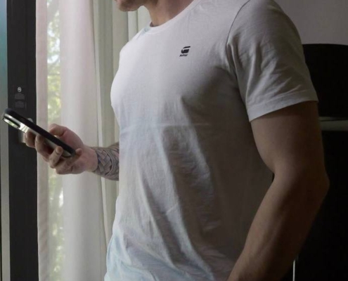 Ein Mann in weißem T-Shirt und Jeans steht an einem sonnenbeschienenen Fenster und lächelt in sein Telefon - Callboy Dennis, eine Hand in der Tasche.