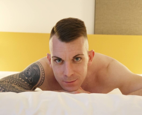 Callboy Dennis, ohne Hemd, mit kurzen braunen Haaren und einer Tätowierung auf dem linken Arm, liegt auf einem Bett und blickt in die Kamera.