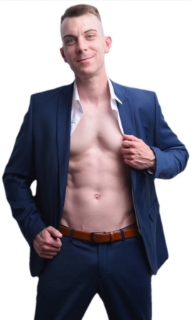 Callboy Dennis, der einen blauen Anzug und ein aufgeknöpftes weißes Hemd trägt, zeigt seine Brust und Bauchmuskeln, während er selbstbewusst vor einem weißen Hintergrund lächelt.