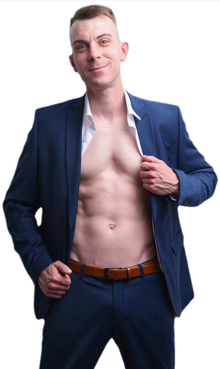 Callboy Dennis, der einen blauen Anzug und ein aufgeknöpftes weißes Hemd trägt, zeigt seine Brust und Bauchmuskeln, während er selbstbewusst vor einem weißen Hintergrund lächelt.