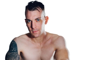 Mann ohne Hemd mit nasser Haut, kurzen dunklen Haaren, blauen Augen und Oberarmtattoo schaut in die Kamera - Callboy Dennis auf weißem Hintergrund.