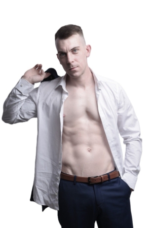 Callboy Dennis, kurzhaarig, posiert vor einem weißen Hintergrund in einem offenen weißen Hemd, blauer Hose mit braunem Gürtel, Jacke über der Schulter.