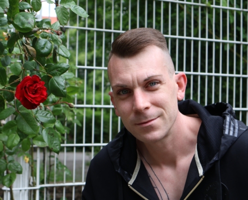 Callboy Dennis steht selbstbewusst vor einem weißen Metallzaun, dessen klare Linien einen Kontrast zur sanften Umgebung bilden. Lässig und entspannt trägt er einen schwarzen Kapuzenpullover, der seine kurzen Haare und hellen Augen, die das natürliche Licht einfangen, perfekt unterstreicht. Neben ihm, auf Zaunhöhe, präsentiert ein Busch seine größte Attraktion – eine einzelne rote Rose in voller Blüte – inmitten leuchtend grünen Laubs, das der Szene Frische und Leben verleiht. Die Gesamtkomposition verbindet urbane Schlichtheit mit dem lebendigen Charme der Natur.