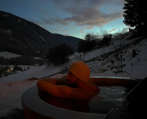 Die Szene fängt eine perfekte Mischung aus Wärme und Kälte ein, als Callboy David inmitten eines Winterwunderlandes im dampfenden Whirlpool Trost findet. Die Strickmütze hält ihn warm gegen die frostige Luft, während er in das wohltuende Wasser eintaucht. Die umliegenden, mit Schnee bestäubten Bäume ragen hoch vor der Kulisse majestätischer Berge empor, deren Gipfel sanft vom schwindenden Licht der Dämmerung berührt werden. Oben blickt eine Mondsichel durch zart verstreute Wolken und verbreitet ein sanftes Leuchten, das die ruhige Atmosphäre dieses Ausflugs verstärkt. Es ist ein Moment, in dem die Schönheit der Natur und die friedliche Einsamkeit zusammenkommen, um Körper und Geist Erholung und Ruhe zu bieten.