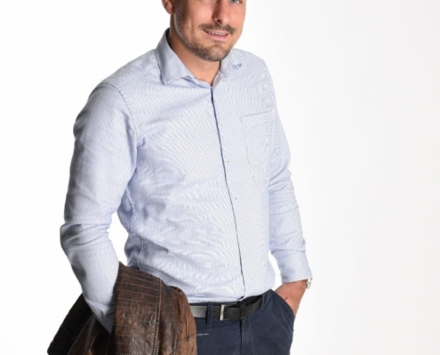 Mann mit kurzem braunem Haar, hellblauem Hemd, dunkler Hose, eine Hand in der Tasche, braune Jacke am Arm, vor weißem Hintergrund.
