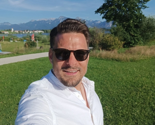 Ein Mann mit Sonnenbrille und weißem Hemd macht ein Selfie auf einer sonnigen Wiese, mit Bäumen, einem Weg und fernen Bergen im Hintergrund.