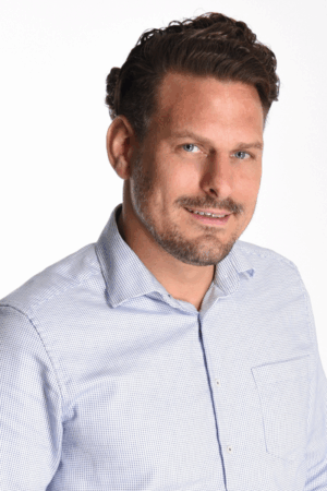 Callboy Simon, mit kurzen, gewellten braunen Haaren und gestutztem Bart, lächelt in einem hellblau karierten Hemd vor einem weißen Hintergrund.