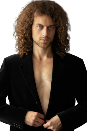 Ein Mann mit langen, lockigen braunen Haaren steht vor einem schlichten weißen Hintergrund. Er trägt einen schwarzen Blazer ohne Hemd darunter und strahlt die selbstbewusste Ausstrahlung von Callboy Anandi aus. Er blickt direkt in die Kamera und ist mitten in der Aktion – beim Zuknöpfen seines Blazers – mit subtiler Anziehungskraft und Selbstsicherheit eingefangen.