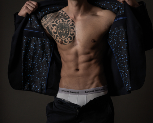 Callboy Dominik, hemdsärmelig in einer dunklen Hose, öffnet einen Blazer, um Tätowierungen auf Brust und Schulter zu zeigen, und posiert selbstbewusst im gedämpften Licht.