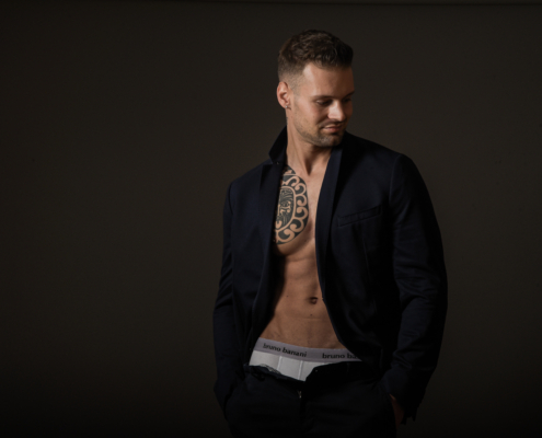 Callboy Dominik, kurzhaarig, steht in einer offenen dunklen Jacke vor einem schwarzen Hintergrund, Brusttattoo und Unterwäscheband sind sichtbar.