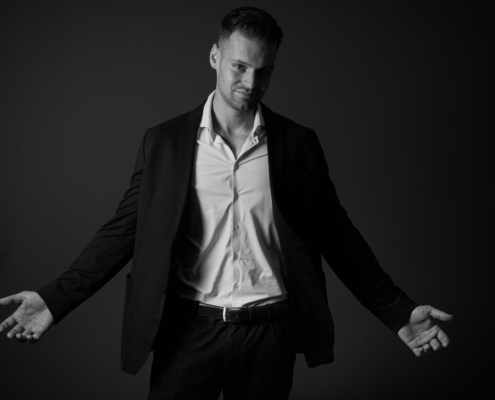 Callboy Dominik steht in diesem dramatischen Schwarz-Weiß-Bild mit ausgebreiteten Armen und erhobenen Handflächen vor einem dunklen Hintergrund und starrt zur Seite.