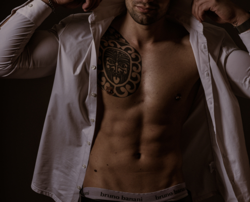Callboy Dominik, ohne Hemd und mit einem Tribal-Tattoo auf der Brust, hebt sein offenes weißes Hemd hoch, um seine Bauchmuskeln zu zeigen; dunkler Hintergrund, schwarze Hose, weiße Unterwäsche.