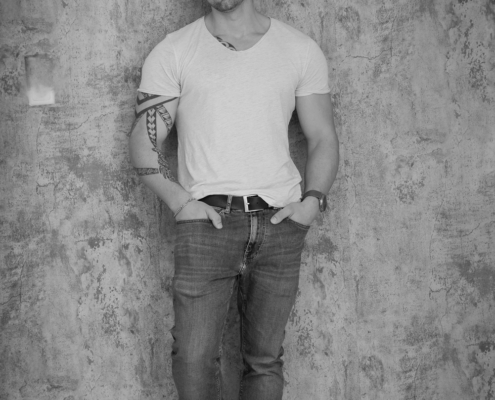 Callboy Dominik, kurzhaarig, lehnt mit den Händen in den Taschen an einer strukturierten Wand, helles T-Shirt, Jeans, Turnschuhe; am linken Arm sind Tätowierungen zu sehen. SCHWARZWEISS.