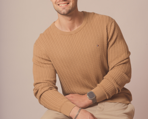 Callboy Dominik, lächelnd, mit kurzen Haaren und gestutztem Bart, sitzt in einem hellbraunen Pullover und einer hellen Hose auf einem Hocker, dahinter eine schlichte Kulisse.