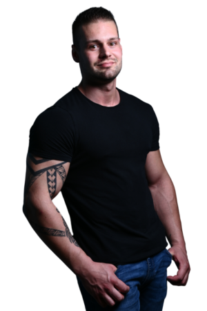 Diese Beschreibung liefert ein klares Bild der Person und der Umgebung. Ein Mann, leger gekleidet in einem schwarzen T-Shirt und Jeans, nimmt eine entspannte Haltung ein und hat die Hände in den Taschen. Auffällig sind seine kurzen Haare und die Tätowierungen auf seinem linken Arm, die seinem Aussehen Details verleihen. Die Szene spielt vor einem schlichten weißen Hintergrund, der die Gesichtszüge und die Kleidung des Mannes ohne Ablenkung hervorhebt. Diese Art von Aufbau hebt in Porträtfotografie oder Werbekontexten häufig Mode oder persönlichen Stil hervor.