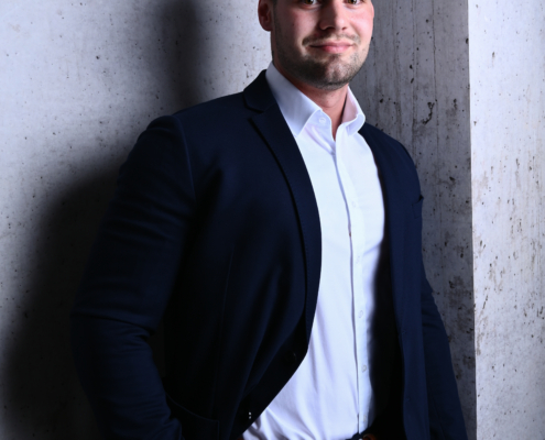 Dieses Bild zeigt eine selbstbewusste Person, die Professionalität und Ausgeglichenheit ausstrahlt. Sein marineblauer Anzug und sein weißes Hemd sorgen für einen klassischen und eleganten Look, der oft mit geschäftlichen oder formellen Anlässen in Verbindung gebracht wird. Die Pose mit einer Hand in der Tasche vermittelt Leichtigkeit und Selbstsicherheit, während sein leichtes Lächeln Zugänglichkeit suggeriert. Der Betonwandhintergrund verleiht dem Foto ein urbanes, zeitgenössisches Flair und macht es für professionelle Porträtfotos, Unternehmensprofile oder persönliche Markenmaterialien geeignet.