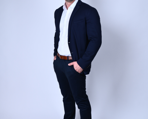 Das Bild zeigt einen selbstbewussten Mann mit gepflegtem Erscheinungsbild. Er trägt klassische, halbformelle Kleidung, bestehend aus einem marineblauen Blazer über einem frischen weißen Hemd, dazu dunkle Hosen und braune Schuhe. Sein kurzes Haar und sein Bart tragen zu seinem gepflegten Aussehen bei. Mit seinen lässig in den Taschen vergrabenen Händen strahlt er ein zugängliches Auftreten aus, während er vor einem schlichten Hintergrund warm in die Kamera lächelt.