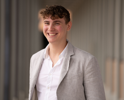 Das Bild zeigt einen fröhlichen und gut gekleideten jungen Mann mit lockigem Haar. Sein hellgrauer Blazer über einem frischen weißen Hemd verleiht ihm ein gepflegtes Aussehen. Der unscharfe Hintergrund mit den tragenden Balken lässt vermuten, dass er sich in einem architektonisch interessanten oder modernen Raum befindet, möglicherweise in Innenräumen. Die Gesamtkomposition unterstreicht das selbstbewusste Auftreten des Mannes vor einem abstrakten Hintergrund.