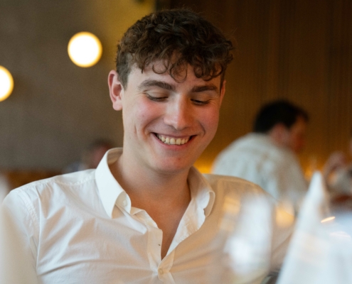 Die von Ihnen beschriebene Szene fängt einen gemütlichen und intimen Moment in einem Restaurant ein. Der Mann mit dem lockigen Haar, der ein weißes Hemd trägt und lächelnd nach unten schaut, lässt vermuten, dass er die Atmosphäre genießt oder vielleicht in einen angenehmen Gedanken oder ein Gespräch vertieft ist. Die warm beleuchtete Umgebung mit verschwommenen runden Lichtern trägt zu einer entspannten und einladenden Atmosphäre bei und verstärkt das allgemeine Gefühl von Wärme und Komfort. Die Anwesenheit anderer Personen im Hintergrund weist auf eine lebhafte und dennoch intime Atmosphäre hin, wie sie typisch für beliebte Restaurants ist, in denen sowohl soziale Interaktion als auch persönliches Vergnügen im Vordergrund stehen.