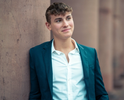 Die Szene, die Sie beschreiben, ruft ein Gefühl klassischer Eleganz und Introspektion hervor. Die Kleidung der jungen Person mit dem dunklen Blazer über einem weißen Hemd verleiht ihr einen zeitlosen und eleganten Look. Ihr kurzes, lockiges Haar trägt zu ihrem markanten Erscheinungsbild bei, während ihr nachdenklicher Ausdruck Kontemplation oder tiefes Nachdenken suggeriert. Die verschwommenen Säulen im Hintergrund tragen zu einem Gefühl von Tiefe und Fokus auf die Person bei und heben sie als Mittelpunkt des Bildes hervor. Diese Komposition könnte Themen wie Reflexion, Kultiviertheit oder sogar Einsamkeit inmitten von Erhabenheit vermitteln.