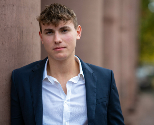 Das von Ihnen beschriebene Foto scheint einen jungen Mann zu zeigen, der einen schicken marineblauen Anzug und ein klassisches weißes Hemd trägt und ein Gefühl von Kultiviertheit und Professionalität ausstrahlt. Die Steinmauer, an der er lehnt, verleiht der Szene Struktur und Kontrast, während der unscharfe Hintergrund mit Andeutungen von Bäumen eine Umgebung im Freien suggeriert, was die ruhige und gelassene Stimmung des Bildes wahrscheinlich noch verstärkt. Sein direkter Blick in die Kamera könnte, je nach Gesichtsausdruck, Selbstvertrauen oder Introspektion vermitteln.