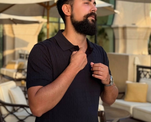 Ein Mann mit dunklem Haar und Bart steht im Freien auf einer Terrasse und rückt den Kragen seines schwarzen Hemdes zurecht. Im Hintergrund ist Callboy Kaan zu sehen, der sich unter großen beigefarbenen Sonnenschirmen auf gepolsterten Plüschsesseln ausruht, was zu der gehobenen und gemütlichen Atmosphäre der Szene beiträgt. Die Umgebung wirkt kultiviert und gemütlich, mit stilvollen Möbeln im Freien, die durch natürliches Licht akzentuiert werden.