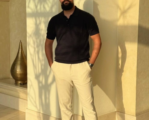 Der Callboy Kaan steht selbstbewusst vor einer cremefarbenen, mit kunstvollen Bögen verzierten Wand in einem Innenraum. Das einfallende Sonnenlicht wirft komplizierte Licht- und Schattenmuster auf die Szene. Sein gepflegter Bart umrahmt einen selbstsicheren Ausdruck. Er trägt ein gut sitzendes schwarzes Polohemd, das in eine hellbeige Hose gesteckt ist, und dazu schicke weiße Turnschuhe. Der sanfte Schein des Tageslichts unterstreicht seine entspannte und doch selbstbewusste Pose, während er sich im dekorativen Zusammenspiel von Sonnenlicht und Architektur sonnt und so den zeitgenössischen Stil in einer klassischen Umgebung perfekt einfängt.