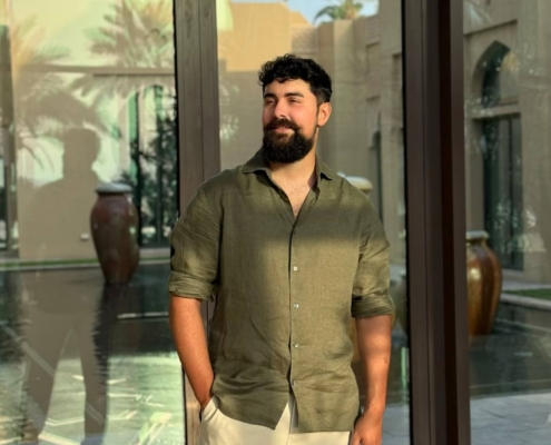 Callboy Kaan steht selbstbewusst vor den großen Glasfenstern, durch die das Sonnenlicht schillernde Muster in den Raum wirft. Sein dunkles Haar und sein gepflegter Bart umrahmen einen nachdenklichen Gesichtsausdruck, der durch ein hellolivfarbenes Hemd ergänzt wird, das ganz natürlich über seinen Körperbau fällt. Das Hemd ist oben leicht aufgeknöpft, was seinem polierten Look ein lässiges Flair verleiht. Dazu trägt er eine beigefarbene Hose, deren klare Linien seinen lässig-eleganten Stil unterstreichen. Hinter ihm schimmern die Reflexionen der Palmen auf der Fensterfläche; das Spiel des Sonnenlichts mit den Blattsilhouetten schafft ein luftiges Ambiente, das sowohl die Person als auch die Umgebung in Wärme und subtile tropische Stimmung taucht.