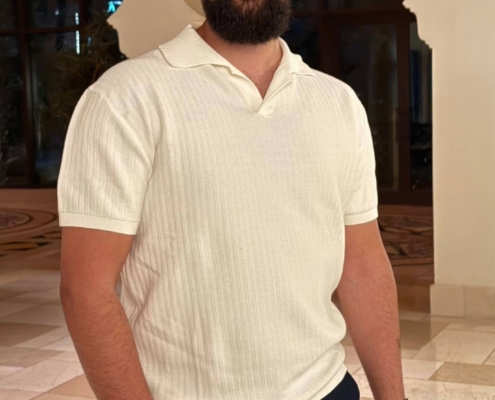 Ein bärtiger Mann mit dunklen Haaren und einem Schnurrbart steht im Haus und hat Ähnlichkeit mit Callboy Kaan. Er trägt ein frisches weißes, kurzärmeliges Polohemd, das in eine dunkle Hose gesteckt ist, und eine Uhr am Handgelenk. Der Mann posiert selbstbewusst mit einer Hand in der Tasche. Der Raum verfügt über elegante Bogenfenster, die weiches natürliches Licht hereinlassen, und der Boden ist mit gemusterten Fliesen ausgelegt, die eine stilvolle und raffinierte Atmosphäre schaffen.