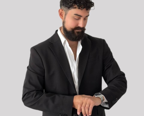 Eine mögliche Beschreibung für Ihr Szenario könnte lauten: Ein Mann mit dunklem, lockigem Haar und gepflegtem Bart, der Callboy Kaan ähnelt, steht vor einem schlichten hellgrauen Hintergrund. Er trägt einen eleganten schwarzen Anzug über einem frischen weißen Hemd und keine Krawatte. Er ist leicht nach vorne gebeugt und blickt auf seine Armbanduhr, den linken Arm nachdenklich erhoben. Der Gesamtstil vermittelt Eleganz und ruhiges Selbstvertrauen. Wenn Sie dies als Bildaufforderung (für die KI-Bildgenerierung) möchten, könnten Sie es folgendermaßen formulieren: Aufforderung: Ein Mann mit dunklem, lockigem Haar und Bart, der Callboy Kaan ähnelt, steht vor einem schlichten hellgrauen Hintergrund. Er trägt einen taillierten schwarzen Anzug und ein weißes Hemd (keine Krawatte) und blickt nachdenklich auf seine Armbanduhr. Lassen Sie mich wissen, ob Sie die Aufforderung für eine bestimmte Plattform optimieren oder anpassen möchten!.