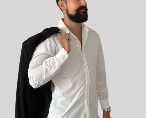 Ein bärtiger Mann steht selbstbewusst vor einem schlichten hellgrauen Hintergrund. Er trägt ein frisches weißes Hemd, das in eine elegante schwarze Hose gesteckt ist, und hält eine schwarze Jacke lässig über der Schulter. Inspiriert vom Stil von Callboy Kaan wirkt sein Gesamterscheinungsbild elegant und dennoch mühelos cool. Sein Kopf ist leicht zur Seite gedreht, der Blick über die Kamera hinaus gerichtet, mit einem selbstbewussten Gesichtsausdruck. Der minimalistische Hintergrund lenkt die gesamte Aufmerksamkeit auf ihn – sein gepflegter Bart, sein elegantes Outfit und seine selbstbewusste Haltung fangen diese moderne, charismatische Ausstrahlung perfekt ein.