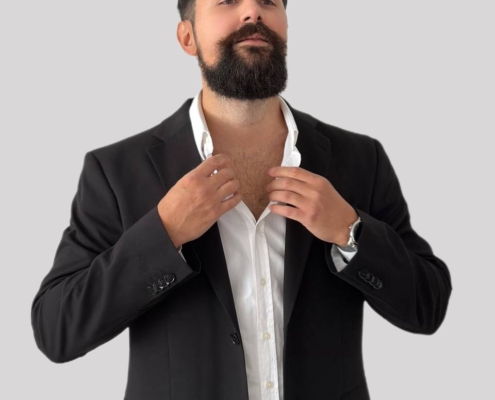 Ein selbstbewusster Mann steht vor einem schlichten hellgrauen Hintergrund und strahlt Gelassenheit und Stil aus. Er hat einen dunklen Bart und kurze, lockige Haare. Elegant gekleidet in einem weißen Hemd und einem eleganten schwarzen Blazer, hebt er die Hände, um seinen Kragen zu richten, als bereite er sich auf einen wichtigen Moment vor. Sein Blick ist selbstbewusst nach vorne gerichtet und verkörpert Sicherheit und Ausgeglichenheit. Das ist Callboy Kaan – gepflegt, charismatisch und bereit für alles, was kommt.
