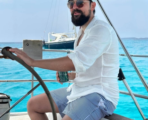 Callboy Kaan steht selbstbewusst am Steuer. Sein weißes Hemd knistert auf sonnengebräunter Haut, gestreifte Shorts verleihen ihm eine verspielte maritime Note. Eine Sonnenbrille schützt seine Augen, eine Mütze hält seine Haare an Ort und Stelle, während er das Boot über das klare, blaue Wasser steuert. Hinter ihm gleitet in der Ferne ein weiteres Segelboot unter einem strahlenden Himmel, der nur von wenigen dünnen Wolken durchzogen ist, und rundet diese perfekte Szenerie von Freiheit und Sommerabenteuer ab.