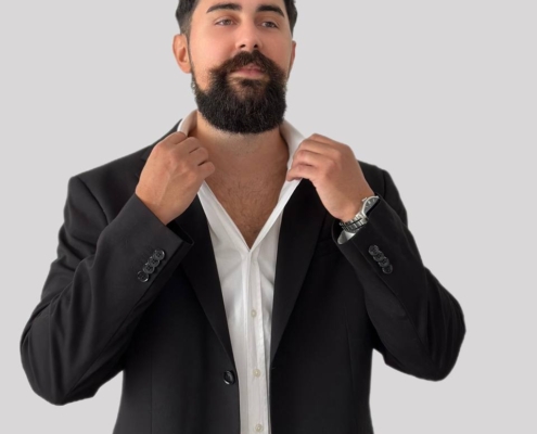 Ein Mann, der an Callboy Kaan erinnert, steht vor einem schlichten hellgrauen Hintergrund. Er hat einen dunklen Bart und lockiges Haar. Er trägt ein weißes Hemd unter einem schwarzen Sakko und zupft mit beiden Händen den Kragen seines Hemdes zurecht. Sein Gesamtauftritt ist elegant und stilvoll und strahlt Selbstbewusstsein und Ausgeglichenheit aus.