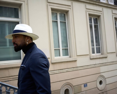 Callboy Hideo steht gelassen und selbstbewusst da. Sein weißer Fedora-Hut wirft einen dezenten Schatten auf seine markanten Gesichtszüge. In seinem dunklen Anzug und einem eleganten schwarzen Rollkragenpullover verkörpert er schlichte Eleganz. Im Hintergrund steht ein reich verziertes beiges Gebäude: Hohe Fenster säumen die Fassade, eingerahmt von dekorativen Stuckarbeiten und klassischen Details, die den Charme der alten Welt andeuten. Sanftes Tageslicht fällt auf die Szene und hebt sowohl Hideos elegante Kleidung als auch die architektonische Raffinesse hinter ihm hervor und verbindet moderne Ausgeglichenheit mit Vintage-Charme.