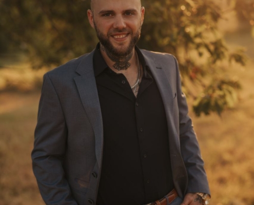 Ein bärtiger Mann mit sichtbaren Nackentattoos steht im Freien im Sonnenlicht. Er trägt einen scharfen blauen Anzug über einem schwarzen Hemd, hat die Hände lässig in den Taschen und schenkt der Kamera ein warmes Lächeln. Hinter ihm sind üppige Bäume und grünes Gras zu sehen, was auf einen Park oder einen in natürliches Licht getauchten Garten hindeutet.