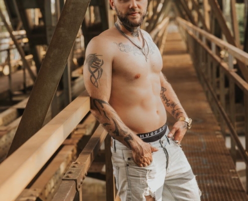 Ein hemdsärmeliger, tätowierter Mann steht selbstbewusst auf einem Metallsteg in einer scheinbar industriellen Umgebung. Er trägt zerrissene Jeansshorts und weiße Turnschuhe. Er lehnt sich lässig gegen das Metallgeländer und blickt in die Kamera, sein direkter Blick strahlt Selbstbewusstsein aus. Der Hintergrund besteht aus industriellen Elementen wie freiliegenden Rohren und Stahlkonstruktionen, die einen düsteren Kontrast zu seiner entspannten Pose und seinem mutigen Stil bilden.