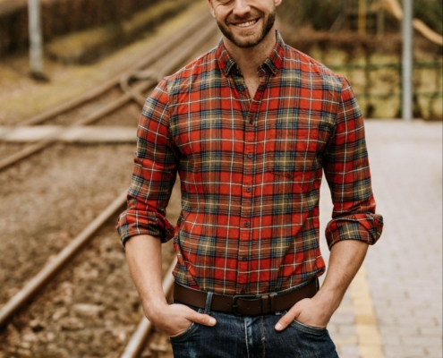 Das Bild zeigt einen Mann, der lässig auf einem Bahnsteig steht. Er trägt ein rotkariertes Hemd und Jeans und hat die Hände bequem in den Taschen verstaut. Sein Gesichtsausdruck ist fröhlich, was darauf schließen lässt, dass er den Moment genießt oder sich auf eine bevorstehende Zugfahrt freut. Hinter ihm erstrecken sich die Gleise in die Ferne und verleihen der Szene Tiefe. Der unscharfe Parkbereich im Hintergrund deutet auf Bewegung oder eine geringe Tiefenschärfe hin, was hilft, die Aufmerksamkeit auf ihn zu lenken und gleichzeitig auf eine malerische Umgebung rund um den Bahnhof hindeutet.