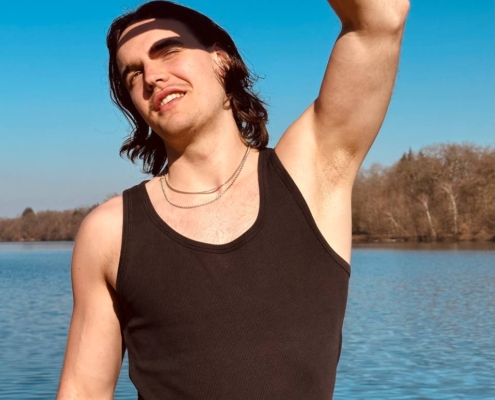 Die Szene wirkt heiter und lebendig und fängt einen Moment der Entspannung und des Naturgenusses ein. Der klare blaue Himmel und die Bäume bilden eine malerische Kulisse für die Person am See. Ihre Kleidung, bestehend aus schwarzem Tanktop und Halsketten, verleiht dieser friedlichen Umgebung einen Hauch von persönlichem Stil. Der Schutz der Augen vor der grellen Sonne lässt auf einen sonnigen Tag schließen, perfekt, um die Natur zu genießen. Diese Beschreibung zeichnet ein idyllisches Bild von Entspannung und Verbundenheit mit der Natur.